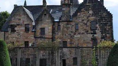 A weathered stone house with a rugged silhouette surrounded by greenery under a cloudy sky.