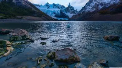 A tranquil lake surrounded by rocky shores and snow-capped mountains under a moody sky.
